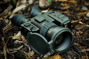 Tactical TBX thermal imaging binocular with control buttons and lens caps in forest ground setting